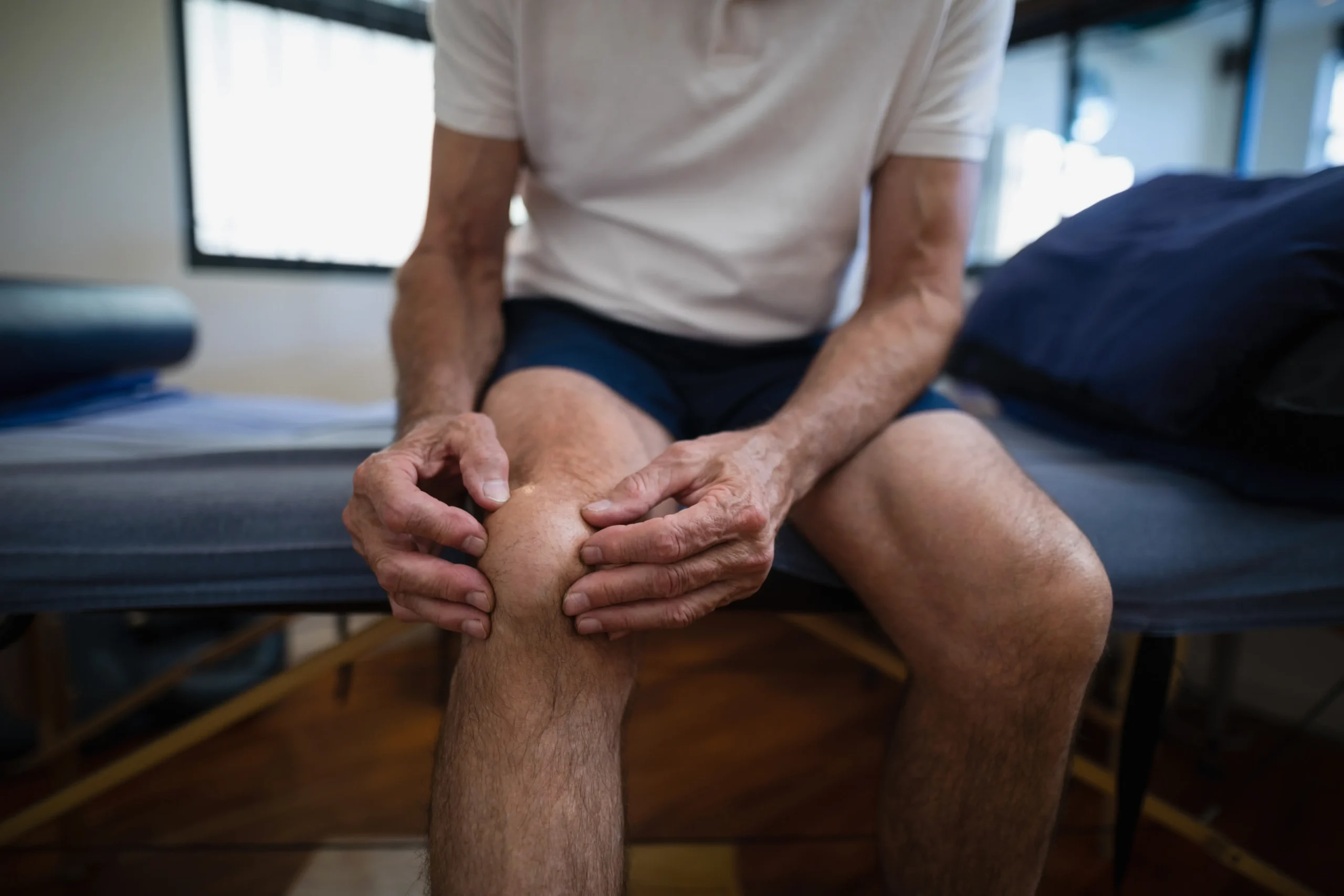 A male athlete holding his knee to express discomfort while sitting on a physical therapist's exam table
